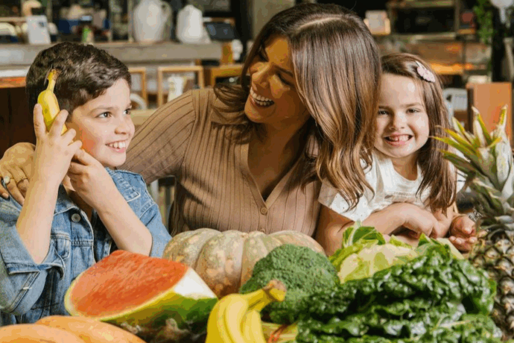 A woman and two children smile together in front of a table filled with colorful fruits and vegetables.