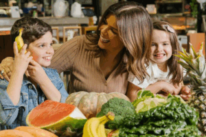 A woman and two children smile together in front of a table filled with colorful fruits and vegetables.