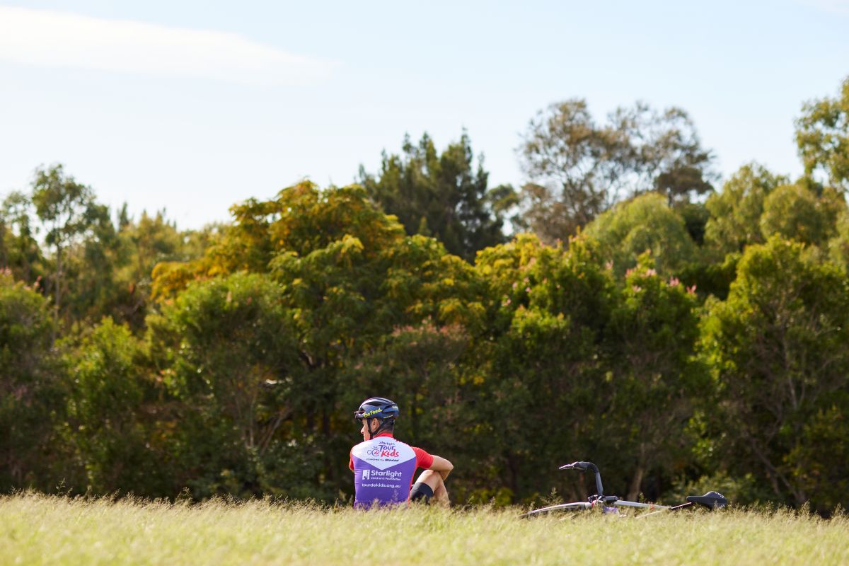 A man in a purple shirt stands in a green field, surrounded by tall grass and a clear blue sky.