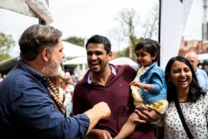 A man and woman with a child enjoying an outdoor event, surrounded by greenery and other attendees.