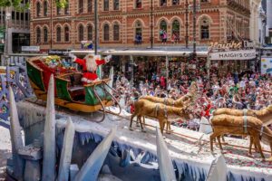 A festive parade float featuring Santa Claus surrounded by reindeer, decorated with colorful lights and holiday ornaments.