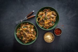 Two bowls of pasta topped with meat and sauce, garnished with herbs, served on a wooden table.