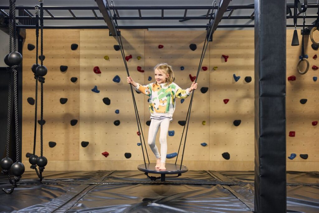 A young girl swings joyfully in a climbing gym, surrounded by colorful climbing walls and safety mats.