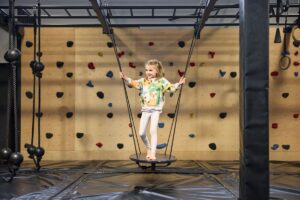 A young girl swings joyfully in a climbing gym, surrounded by colorful climbing walls and safety mats.