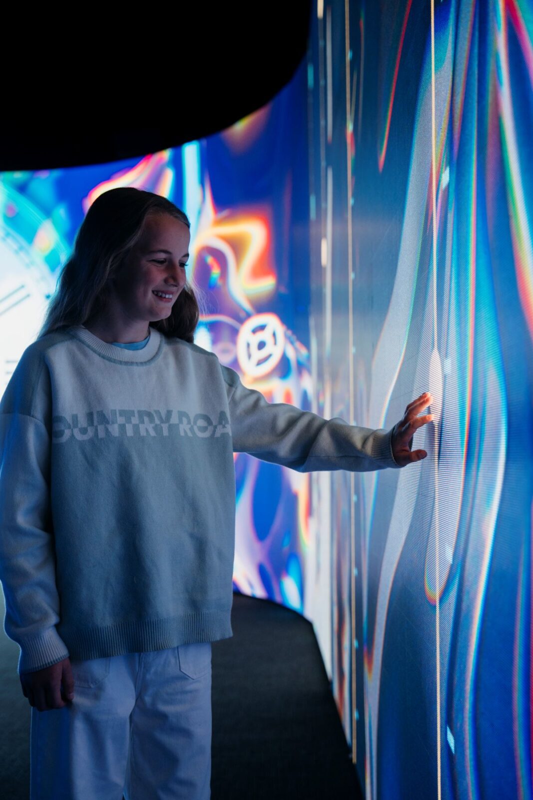 A girl stands before a vibrant wall of colorful lights, creating a striking visual display in the background.