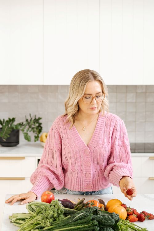 A woman in a pink sweater stands by a table filled with fresh vegetables, smiling at the camera.