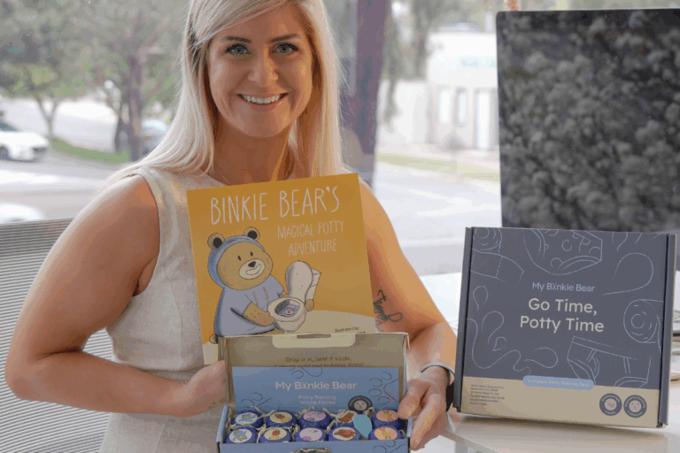 A woman holds a box of books and a teddy bear, smiling as she prepares to share them with someone special.