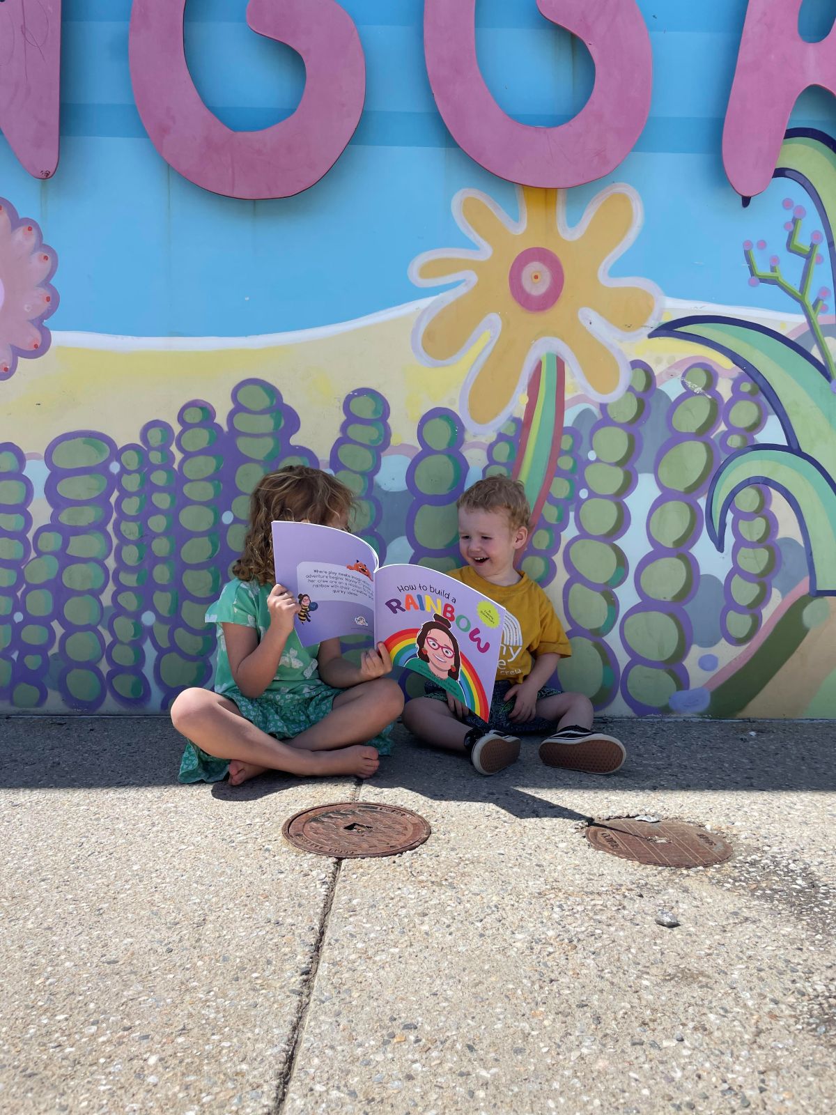 Two children sitting on the ground, engrossed in reading their books, surrounded by a peaceful outdoor setting.