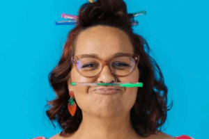 . A woman with a mustache playfully holds a pencil in her mouth, looking thoughtful and engaged in her work.