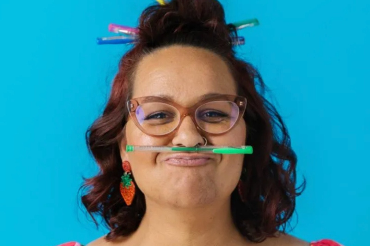 . A woman with a mustache playfully holds a pencil in her mouth, looking thoughtful and engaged in her work.