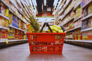 A shopping basket filled with assorted fruits and vegetables in a supermarket aisle.