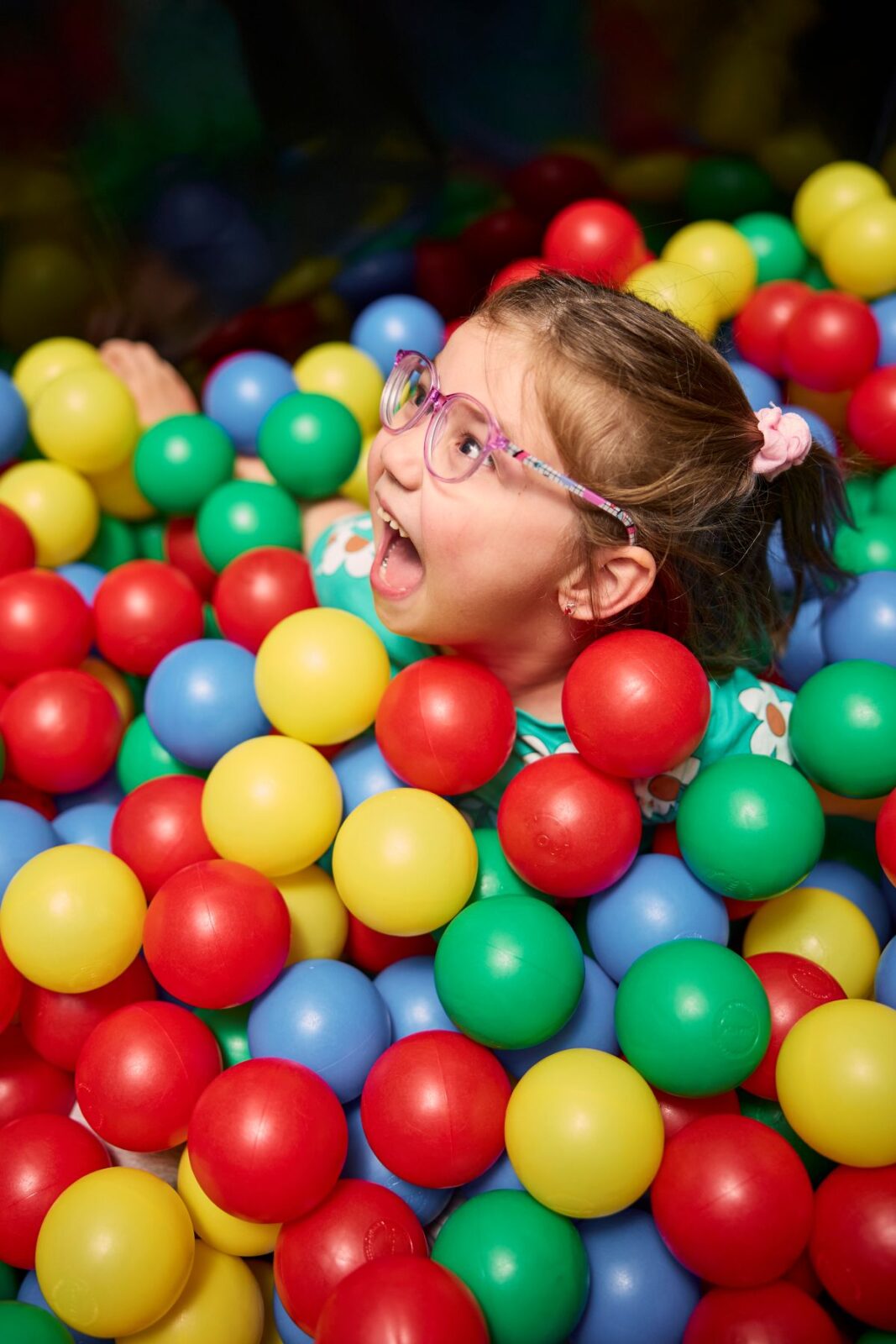 A young girl with glasses joyfully plays in a colorful ball pit filled with various plastic balls.