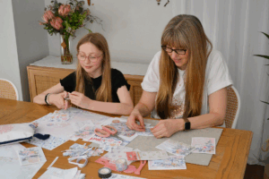 Two women collaborate at a table, creating handmade cards together with various craft supplies spread out.