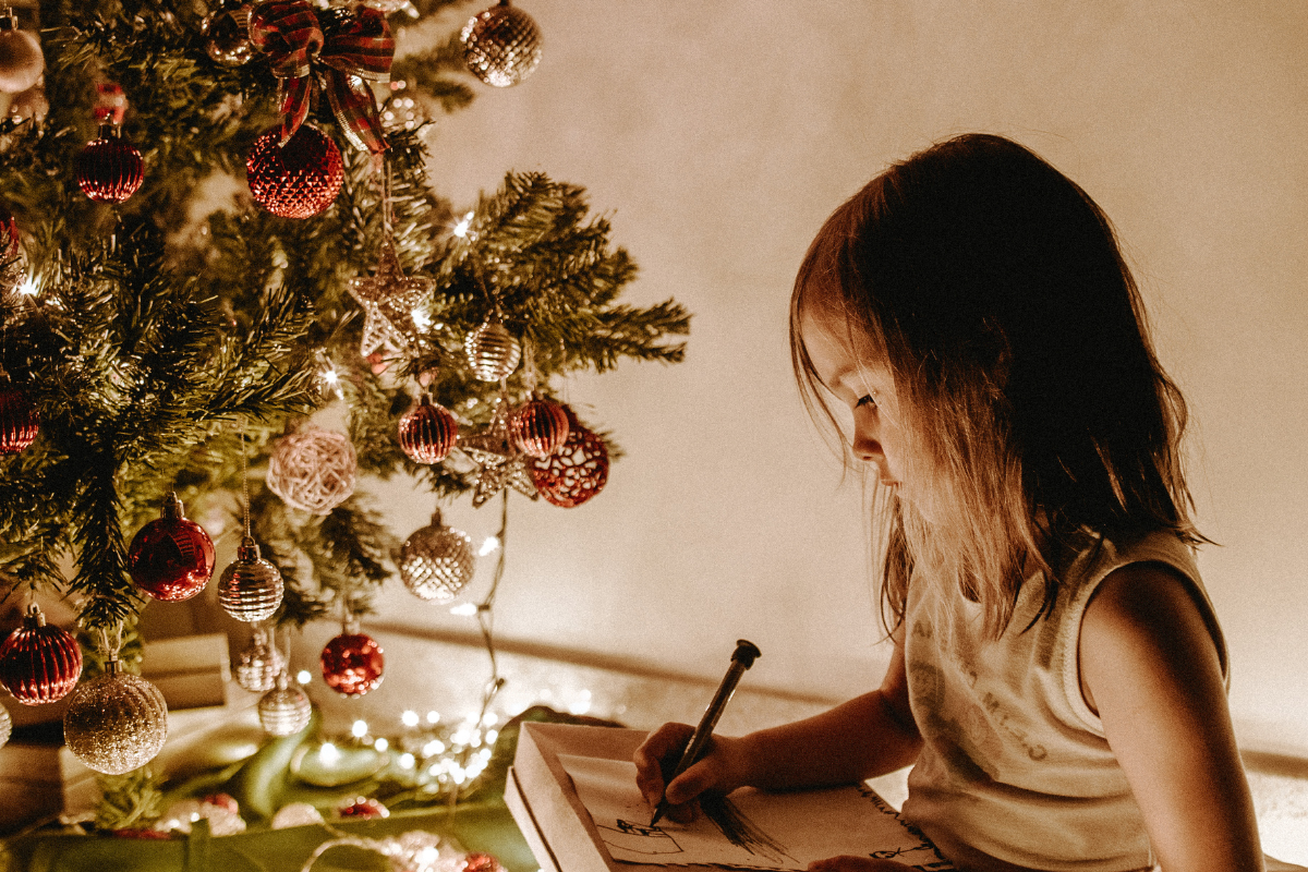 A young girl writes in a book beside a decorated Christmas tree, surrounded by festive lights and ornaments.