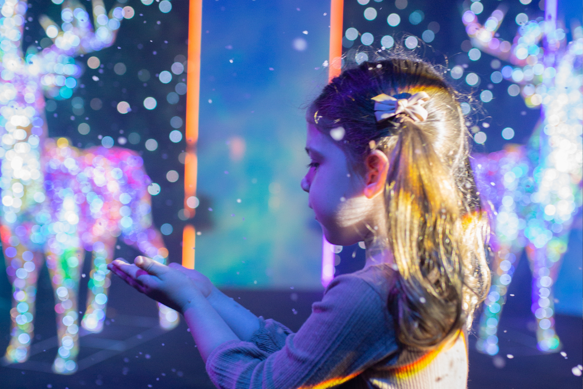 A little girl gazes at a deer during a colorful indoor light show.