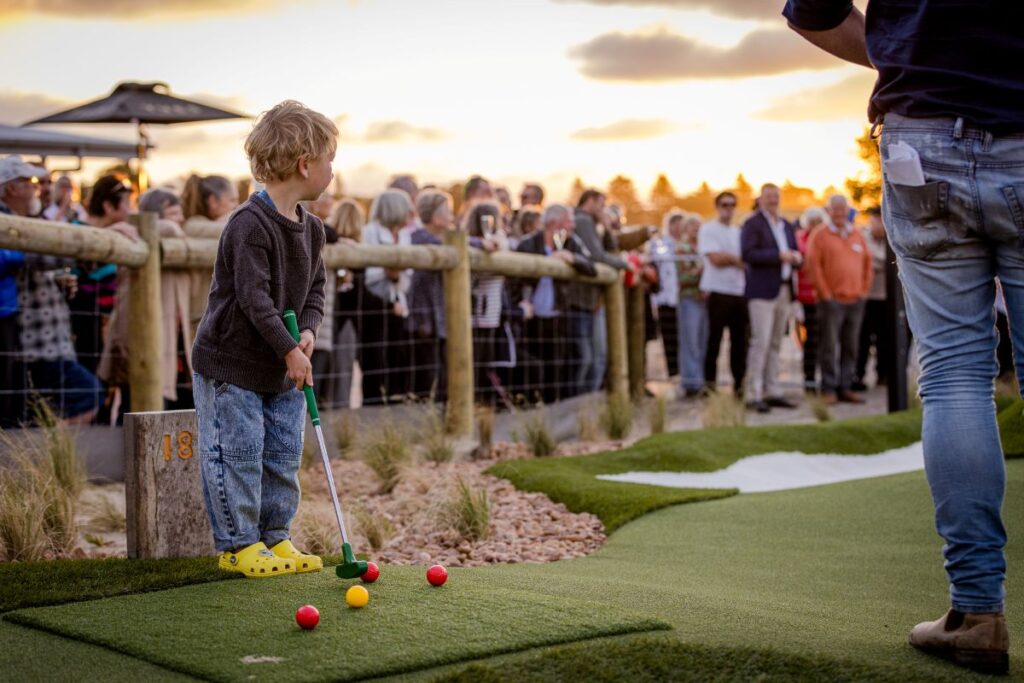 A young boy enjoys playing mini golf as the sun sets, casting a warm glow over the course.
