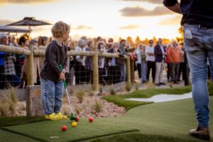 A young boy enjoys playing mini golf as the sun sets, casting a warm glow over the course.