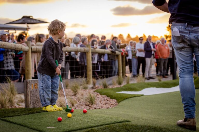 A young boy enjoys playing mini golf as the sun sets, casting a warm glow over the course.