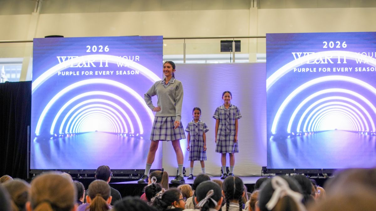 A group of children stands on stage in front of a large screen, smiling and engaging with the audience.