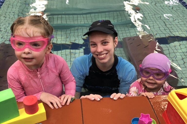 An adult and two children with swim goggles sit on a table by the pool, excitedly preparing for their swimming activities.