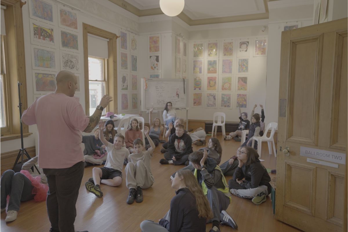 A group of children sitting on the floor, attentively listening to a man speaking in front of them.