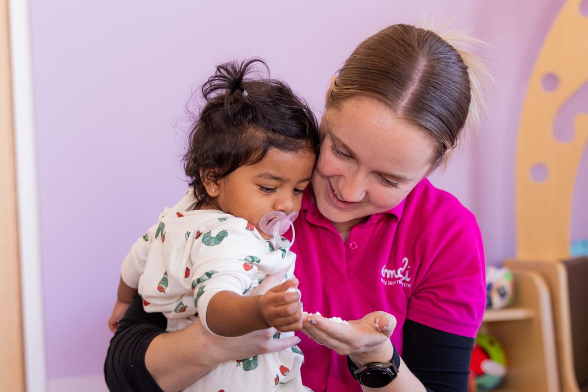 A woman embraces her baby in a cheerful playroom, featuring bright colors and an array of toys.