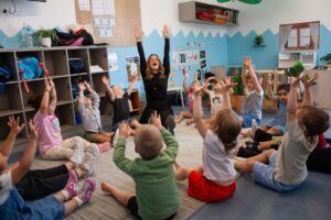 A teacher engages with children in a classroom setting, surrounded by learning resources and student artwork on the walls.