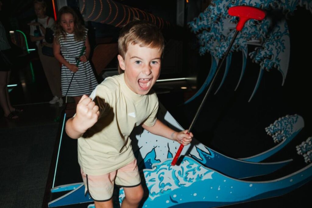 A young boy enjoys playing mini golf at a vibrant theme park, focused on his next shot.