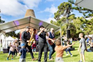 A lively scene of people playing jazz music together on a grassy field, surrounded by nature and children dancing and enjoying the moment.
