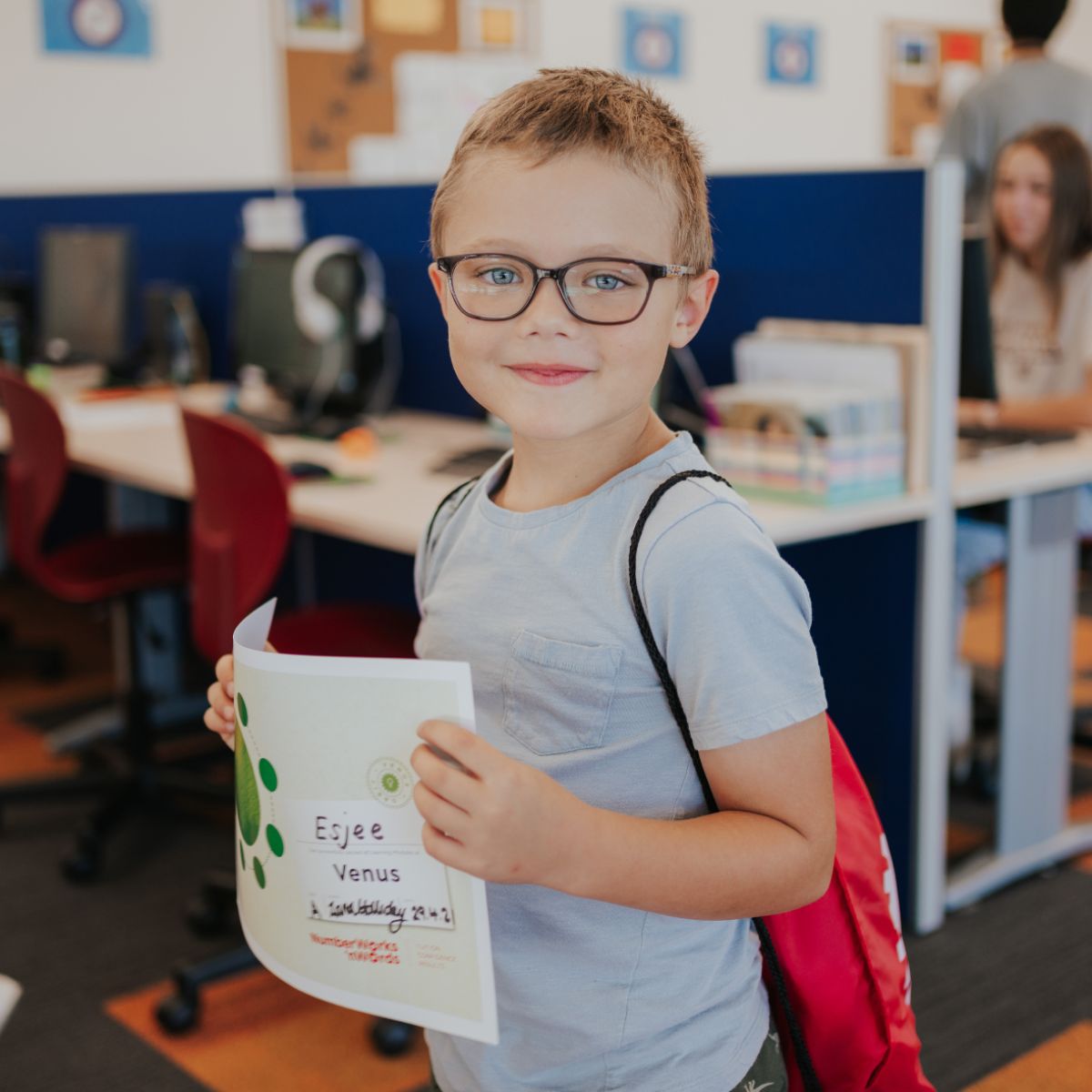 A young boy wearing glasses stands in a classroom proudly holding a certificate of achievement