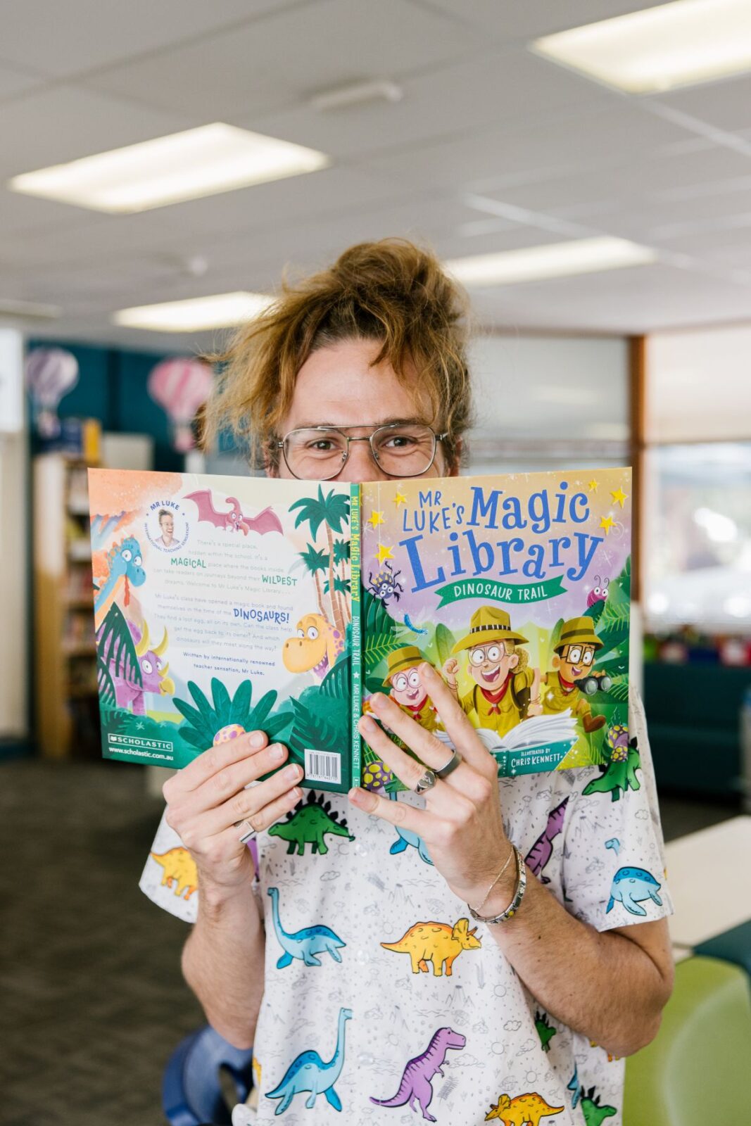 A teacher wearing a dinosaur shirt holds up two books, smiling at the camera in a bright, cheerful setting.