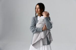 A woman in a grey sweater gently holds a baby in her arms, smiling softly at the camera.