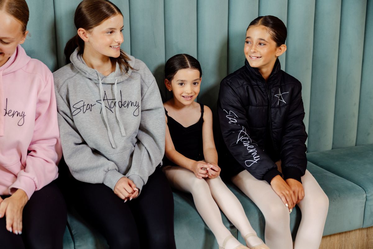 Girls sit on a couch, smiling and showing off their ballet shoes.