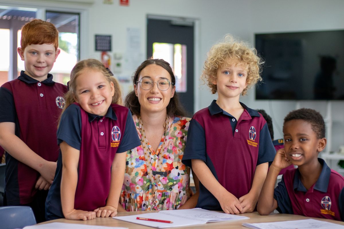 A teacher smiles with her students in a bright classroom filled with educational posters and desks.
