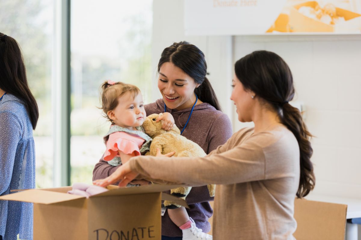 A woman cradles a baby while another woman holds a teddy bear, both smiling in a warm, nurturing setting.