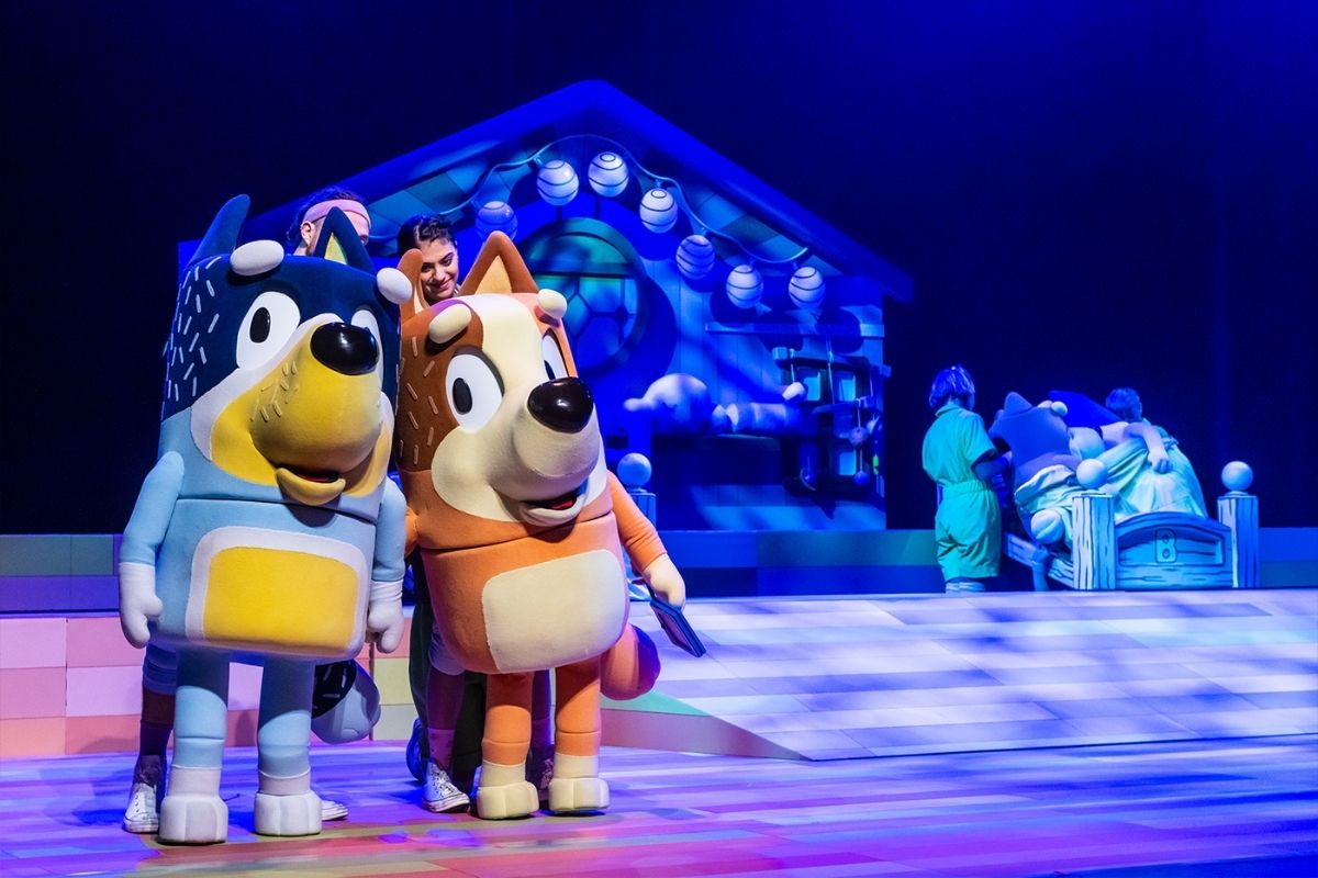 Two cartoon characters stand on stage, smiling and engaging with an audience in a colorful theater setting.