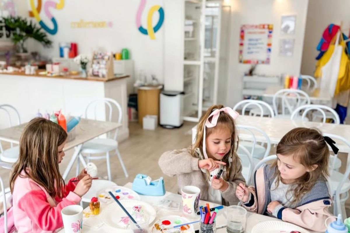 Three young girls are engaged in painting at a table, surrounded by colorful art supplies and their artwork.
