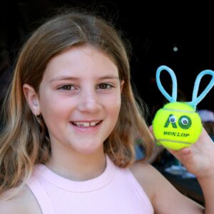 A cheerful young girl grips a tennis ball with bunny ears, showcasing her playful spirit.