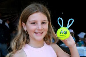 A cheerful young girl grips a tennis ball with bunny ears, showcasing her playful spirit.