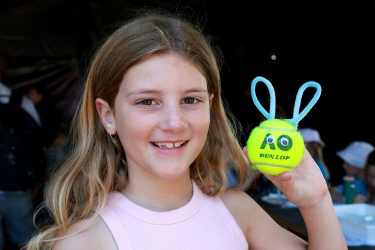 A cheerful young girl grips a tennis ball with bunny ears, showcasing her playful spirit.