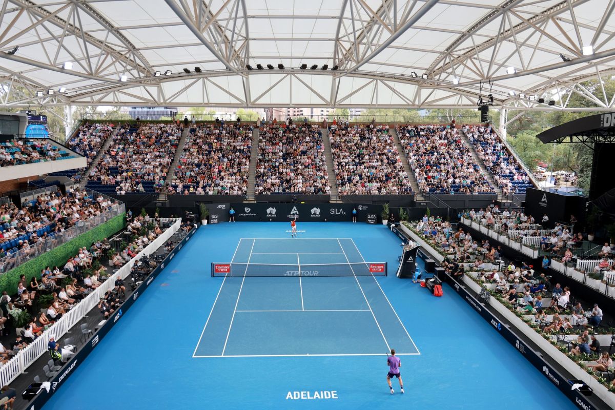 A large crowd attentively watches a tennis match, with players visible on the court in the background.