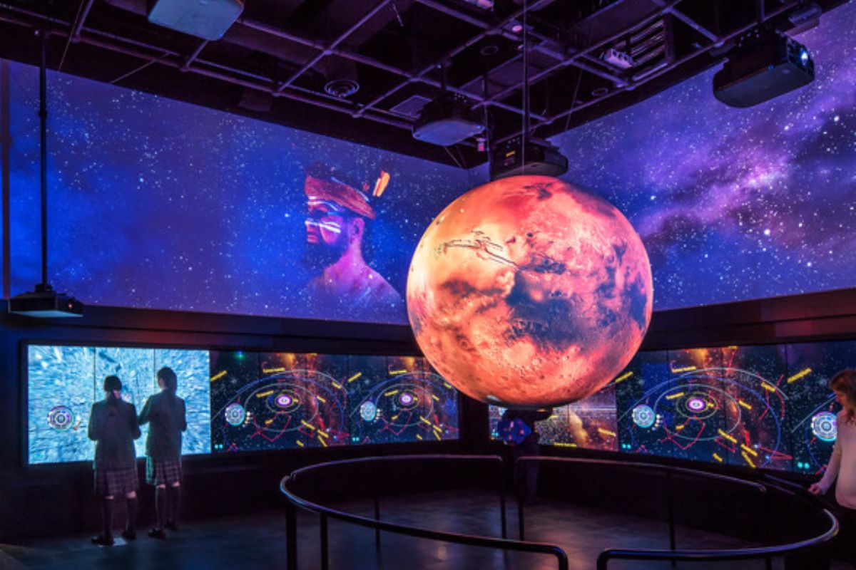 Individuals observe a large screen featuring a vibrant planet, engaged in discussion.