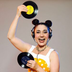 A woman in a polka dot dress smiles while holding a vinyl record in her hands.