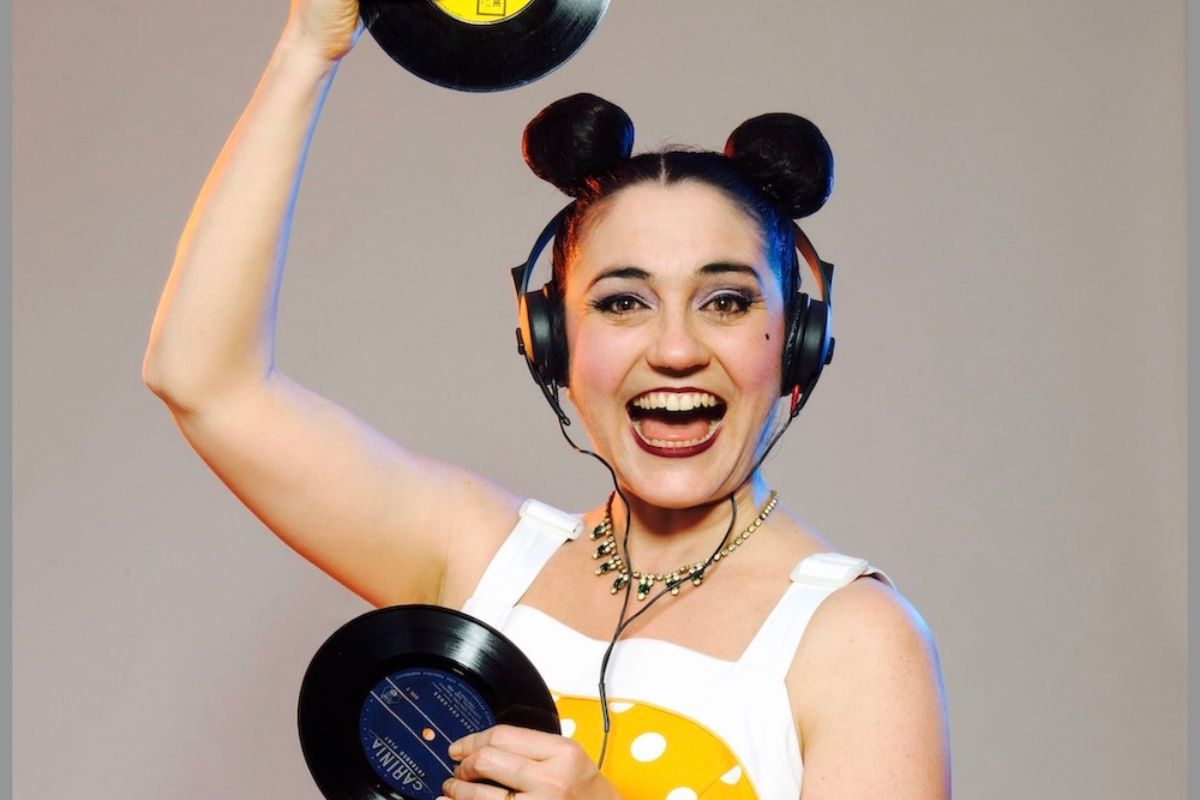 A woman in a polka dot dress smiles while holding a vinyl record in her hands.