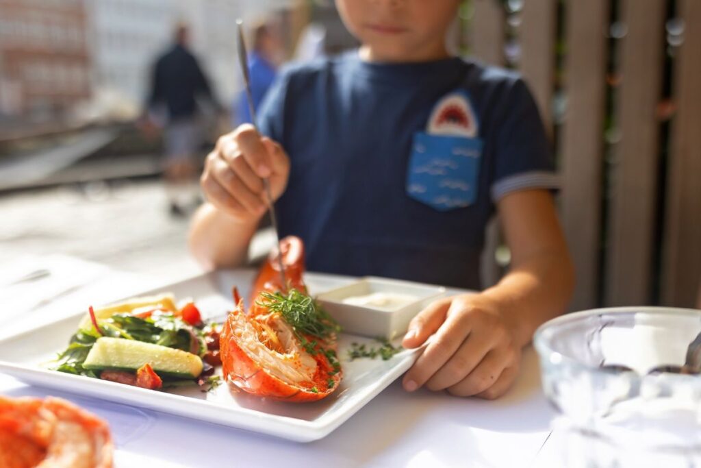 A young boy enjoys a lobster served on a plate, smiling as he takes a bite.