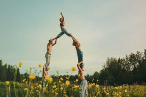 A group of people dressed in denim jeans pose in the shape of a human pyramid in a field of yellow flowers under a cloudy blue sky