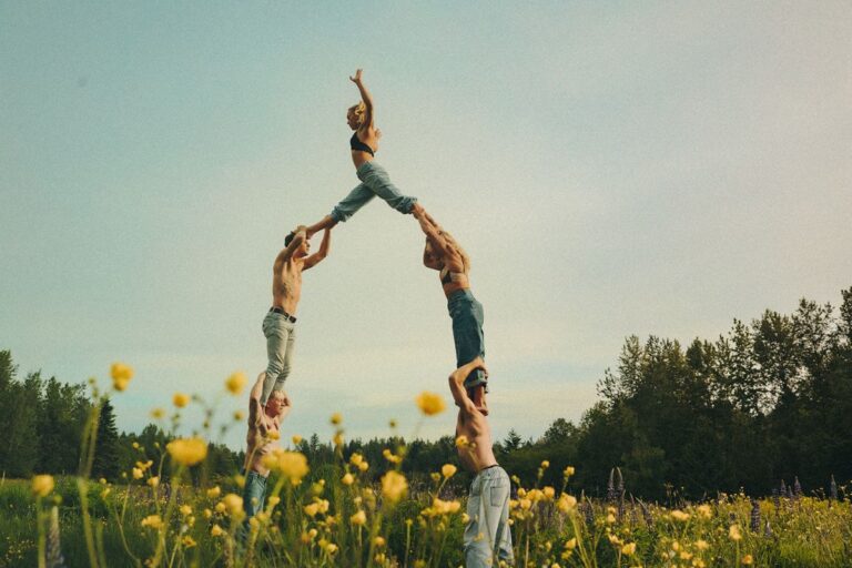 A group of people dressed in denim jeans pose in the shape of a human pyramid in a field of yellow flowers under a cloudy blue sky