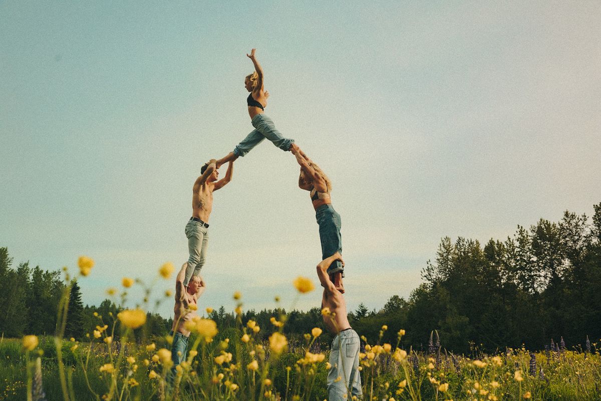 A group of people dressed in denim jeans pose in the shape of a human pyramid in a field of yellow flowers under a cloudy blue sky