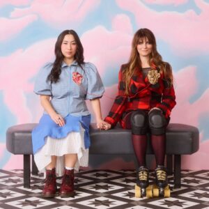 Two women with roller skates sit on a bench, engaged in conversation and laughter in a vibrant environment.