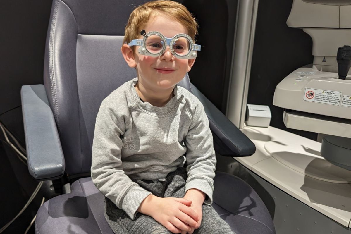 A young boy sitting in an eye exam chair, looking curiously at the eye doctor in a bright examination room.